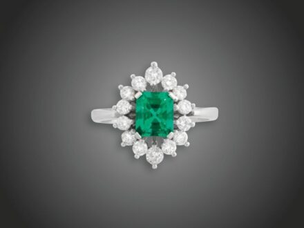 55. tétel, Női gyűrű  Fehérarany 585, karmos foglalatban 14 briliáns cca. 0,76 ct és 1 smaragd cca. 1,50 ct ékítéssel, 5,7 g, átmérő: 17,6 mm, tanúsítvánnyal, smaragd kezelt