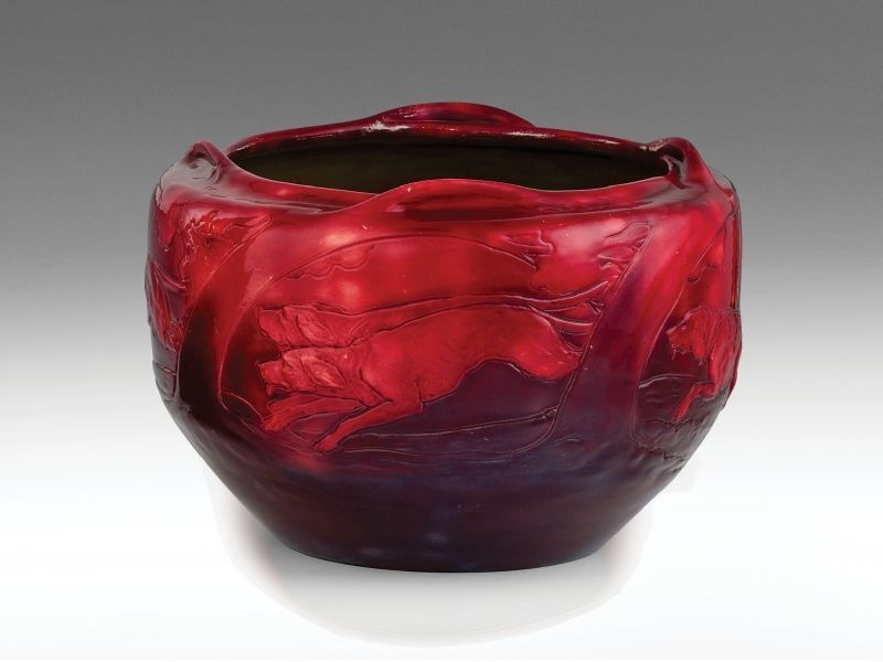 472. tétel, Zsolnay szecessziós virágtartó  Formába öntött porcelánfajansz. Plasztikus  elemekkel díszített, felületén kontúrrajzos,   ill. reliefszerű a laterna magica ötletén alapuló  jelenet: nyulat üldöző vadászkutyák.  Felületén színes eozinmázakkal