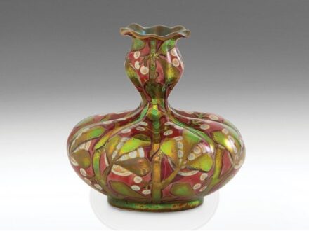 467. tétel, Zsolnay szecessziós kisváza  Formába öntött keménycserép. Gerezdelt felületén fémesfényű, fényes és matt, váltakozó eozinmázakkal festett, részben savmaratott leveles-bogyós (fagyöngy) dekor. Szájperemén apró restaurálás.  Alján jelzett: massz