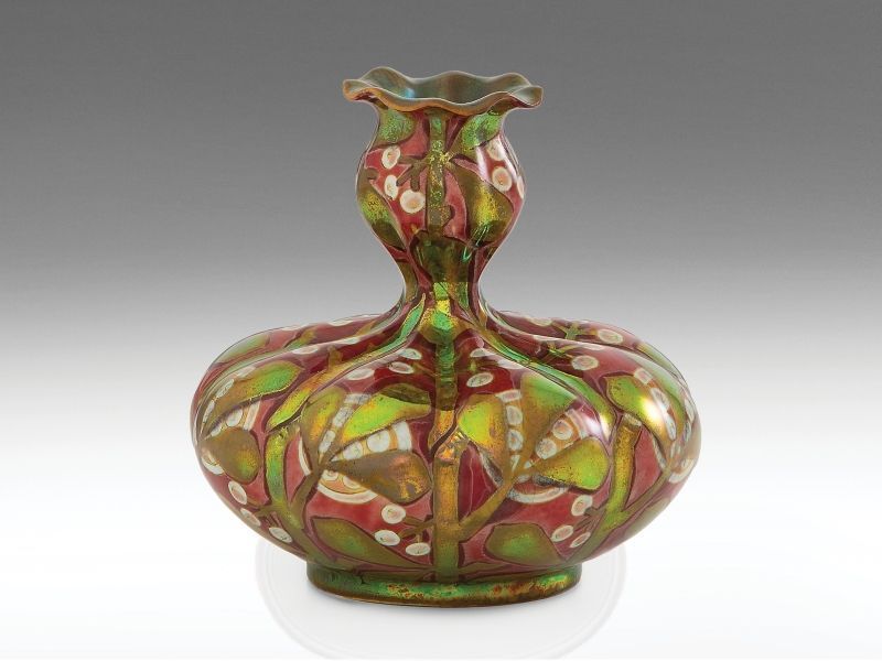 467. tétel, Zsolnay szecessziós kisváza Formába öntött keménycserép. Gerezdelt felületén fémesfényű, fényes és matt, váltakozó eozinmázakkal festett, részben savmaratott leveles-bogyós (fagyöngy) dekor. Szájperemén apró restaurálás. Alján jelzett: massz