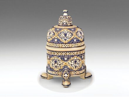 447. tétel, Zsolnay fedeles díszedény – az Arab-sorozatból  Formába öntött porcelánfajansz. Felületén áttört, arabeszk díszítmények színes festéssel és aranyozással. Fedele hajszálrepedt, alján restaurált.   Formaterv: Sikorski Tádé, 1889.  Alján jelzett: