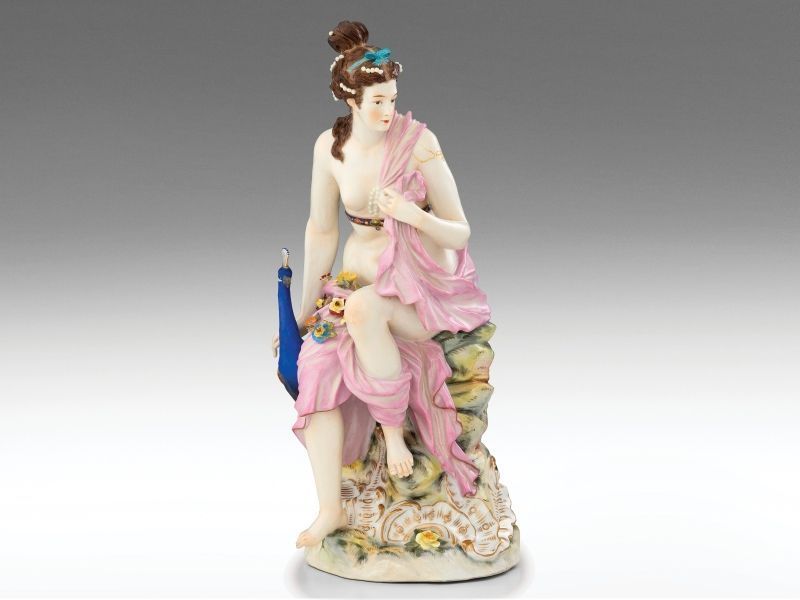 412. tétel, Kisplasztika - Héra, az Olümposz királynője   Színesen festett és aranyozott porcelán. Rokályos talapzaton ülő Héra istennő, oldalához simuló szent madarával, a pávával. Rózsaszín leplén plasztikus virágdíszek. Tűzrepedt, apróbb csorbák, ill.