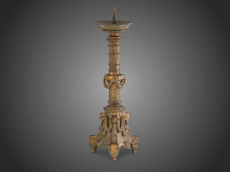 392. tétel, Klasszicista gyertyatartó Öntött, patinázott, aranyozott bronz. Akantuszlevelekkel díszített, osztógyűrűkkel tagolt oszlopon applikált babérfüzérek és rozetták. Aranyozása kopott, talpazatában utólagos szerelékkel. XVIII. sz. második fele.