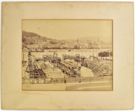 1900 Erzsébet-híd építése, rajta a budai panorámával, a Gellért-hegy részletével, háttérben a Tabán házaival, a tabáni szerb Szent Demeter-templommal, kicsit fentebb pedig az egytornyú Alexandriai Szent Katalin-templom, a pesti oldalon, az építkezést elkerítő kerítésen szecessziós reklám-plakátokkal, nagy, exkluzív méretű keményhátú fotó, paszpartuban, a felületén kis foltokkal, foltos paszartuval, és foltos hátoldallal, 34,5x46,5 cm, paszpartu: 53x69 cm.  Feltehetőleg eredetileg: "A budapesti Erzsébet-híd vasszerkezete. Gyártotta és szerelte az 1898-1903. években a M. Kir. Államvasutak Gépgyára. Bp., [1903], Athenaeum." c. kiadványhoz készült